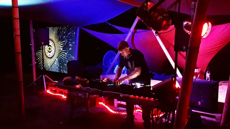 Bring The Beat Mack DJing under a canopy at night, lit red and blue, with visuals on a screen behind