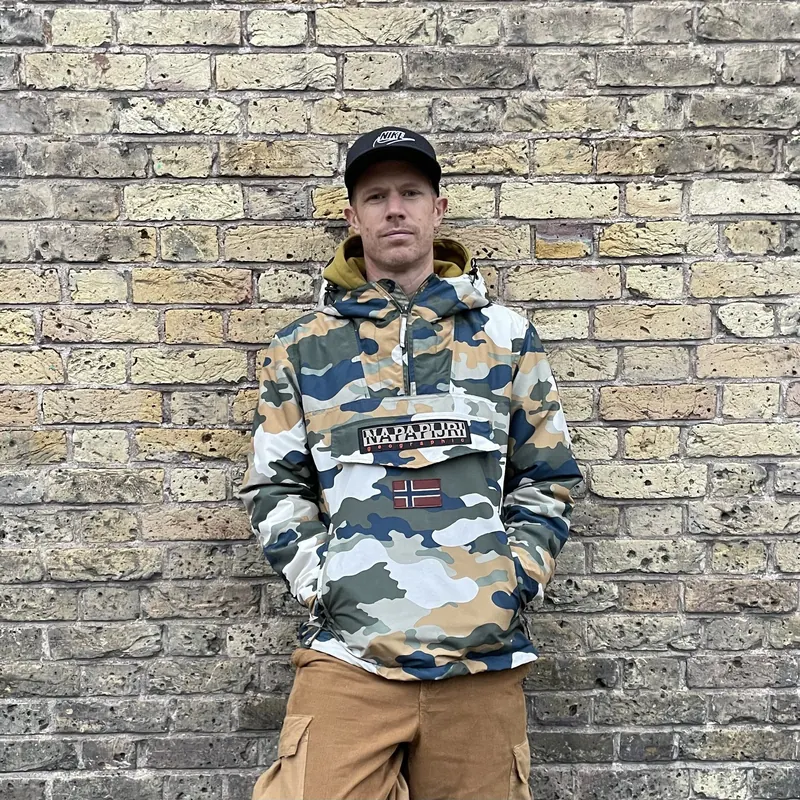 Portrait of Bring The Beat Mack in a camo jacket and cap against a London brick wall