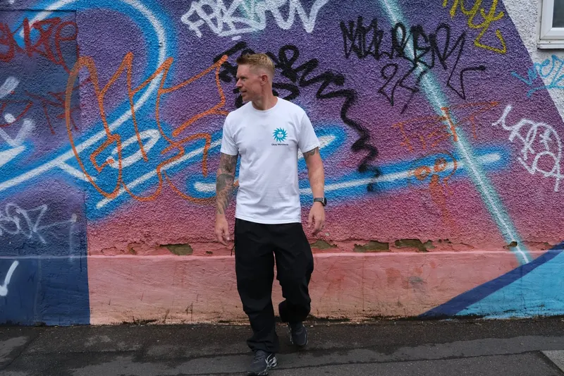 Bring The Beat Mack in a white T-shirt in front of a colourful graffiti wall